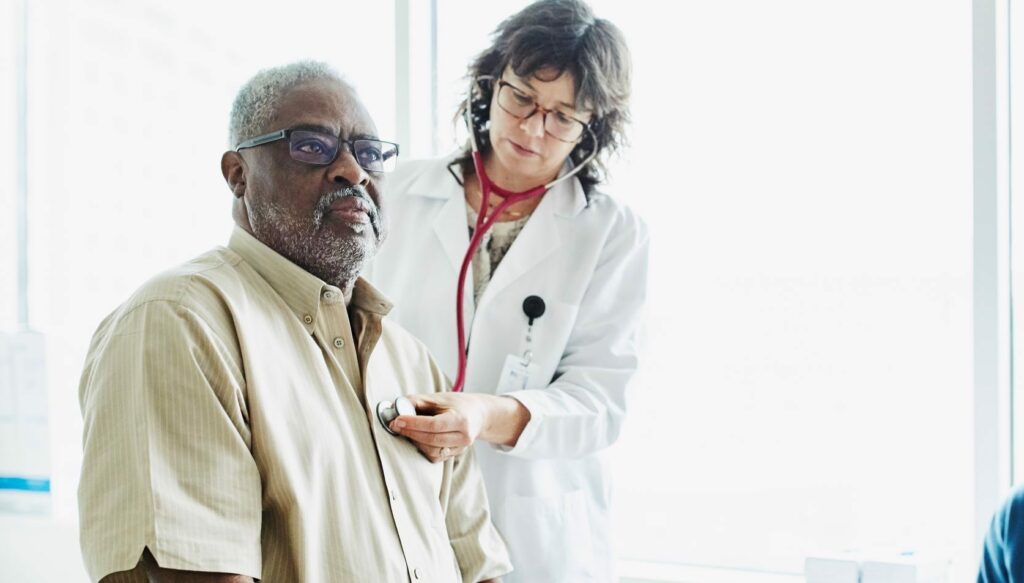 Female doctor listening to an older man's heart while a younger woman watches.