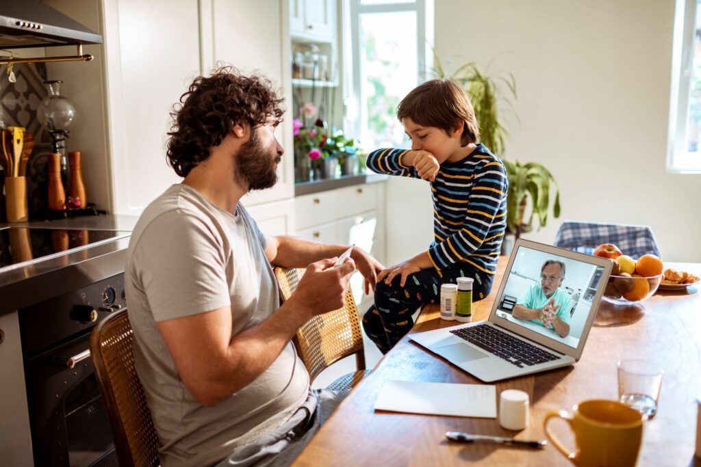 Man sits at table while looking at a boy sitting on the table covering his cough.